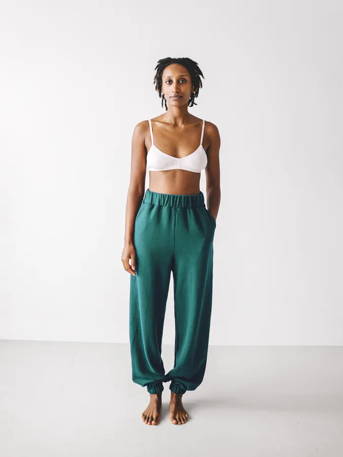 A model in a white bralette and forest-green jogger-style sweatpants with elasticized cuffs, standing barefoot against a white studio background.