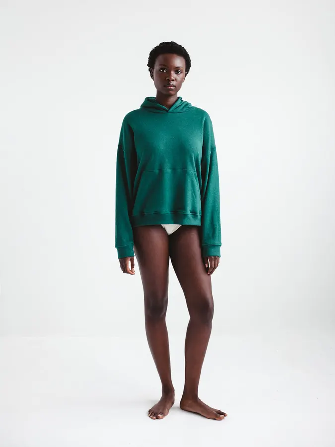 A model wearing a forest-green pullover hoodie with a kangaroo pocket, standing barefoot against a white studio background.