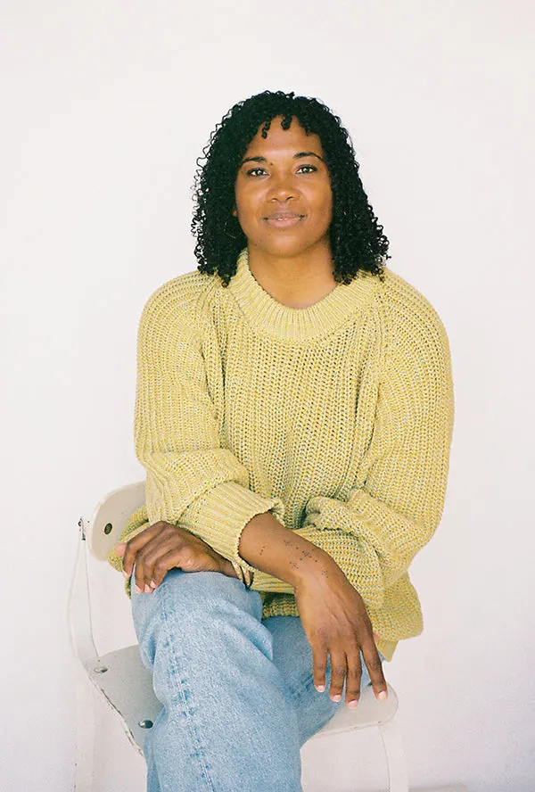 Designer Aliya Wanek seated on a white chair against a plain wall, wearing a chunky yellow knit sweater and blue jeans, smiling at the camera.