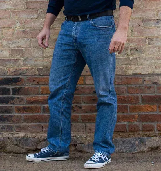 A man's lower body in profile, wearing medium-wash denim jeans, a black belt and navy sneakers, standing against a brick wall.