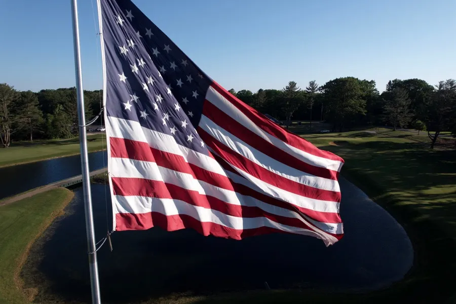A large American flag flies from a pole against a clear blue sky, with a pond and green lawn below.