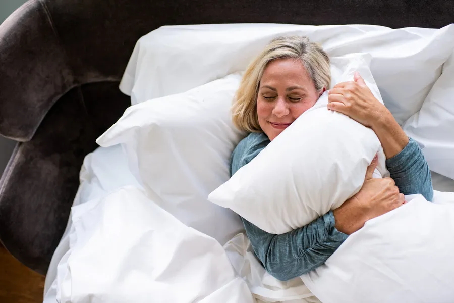 A woman with closed eyes lies in bed hugging a white pillow, surrounded by white sheets.