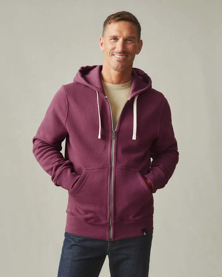 A smiling man wears a maroon zip-up hoodie, hands in the front pockets, against a plain light grey studio background.