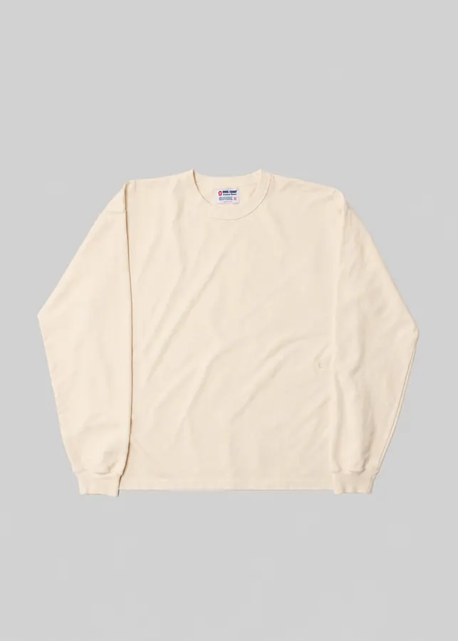 A natural off-white long-sleeve crewneck t-shirt with a woven brand label at the collar, laid flat against a light grey background.