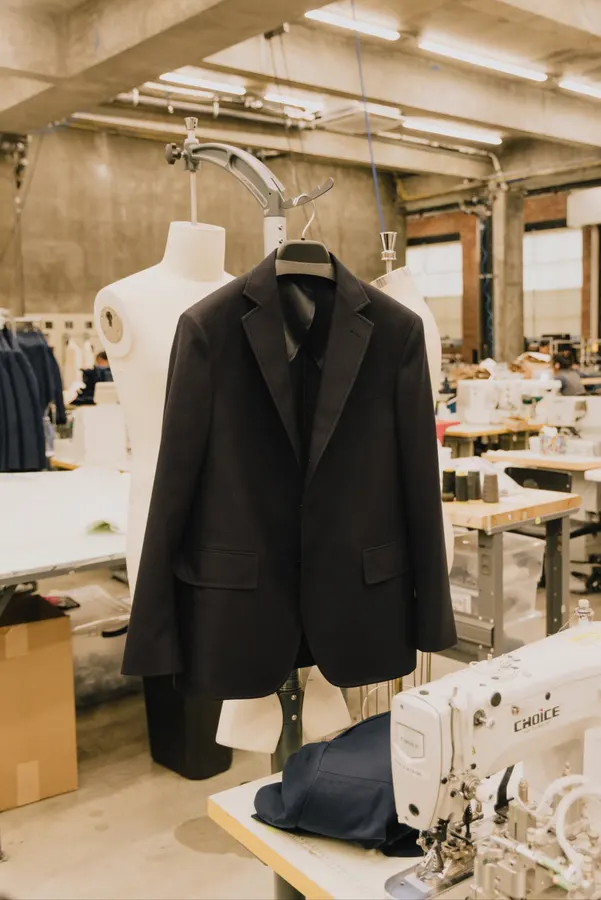 A navy wool blazer hangs on a dress form amid sewing machines, fabric bolts and cardboard boxes on an American Trench factory floor.