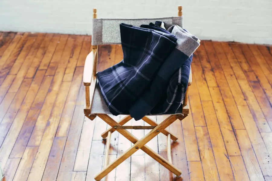 A folded navy and grey plaid wool blanket draped over a wooden director's chair on a hardwood floor against a white brick wall.