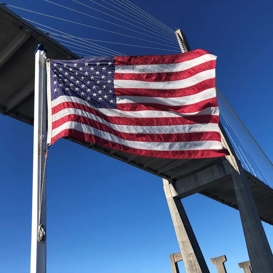 A large American flag flying from a pole beneath a cable-stayed bridge, backlit against a clear blue sky.