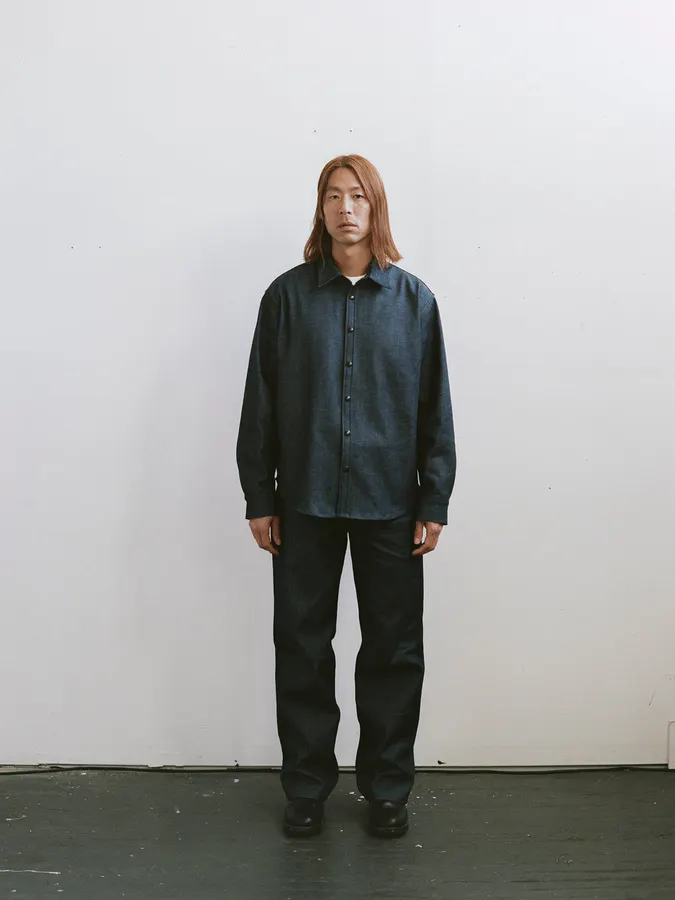 A man stands facing the camera against a plain white studio wall, wearing a dark denim button-up overshirt and black trousers.