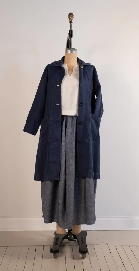 A navy linen swing coat with patch pockets and covered buttons, displayed buttoned over a white blouse and grey linen skirt on a dress form against a plain wall.