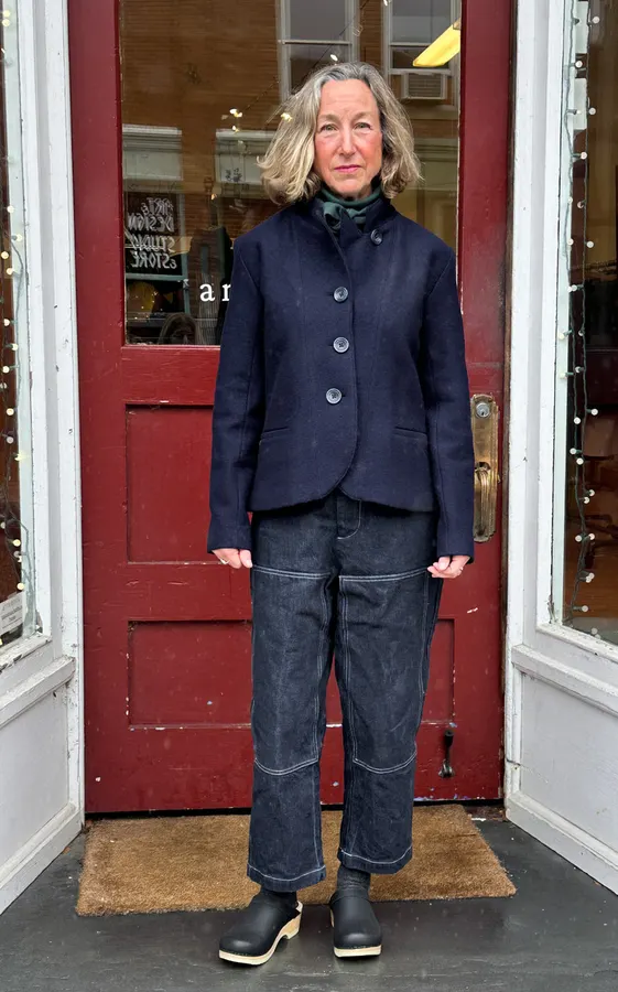 A woman wearing dark indigo wide-leg denim garden pants with contrast topstitching, a navy jacket, and clogs, standing in front of a red shopfront door.