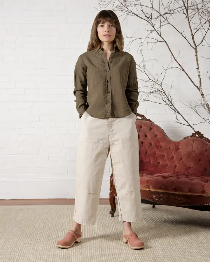A woman with shoulder-length brown hair wears an olive linen button-front shirt with cream wide-leg garden pants and pink suede clogs, standing in front of a tufted red antique settee against a white brick wall with a bare birch branch.
