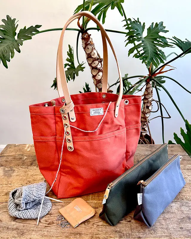 A red waxed-canvas tote bag with leather handles, a tan leather wallet, and two zippered canvas pouches arranged on a wood table in front of a large houseplant.