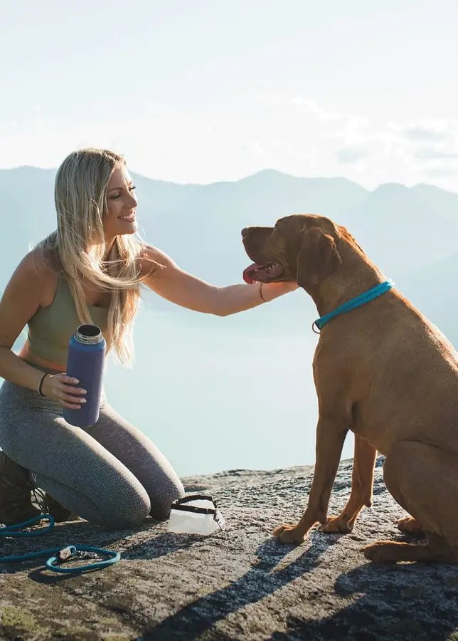 A woman kneeling on a mountain overlook pets a large brown dog wearing a teal rope collar, with a lake and mountains in the background and a matching teal leash coiled on the rock.