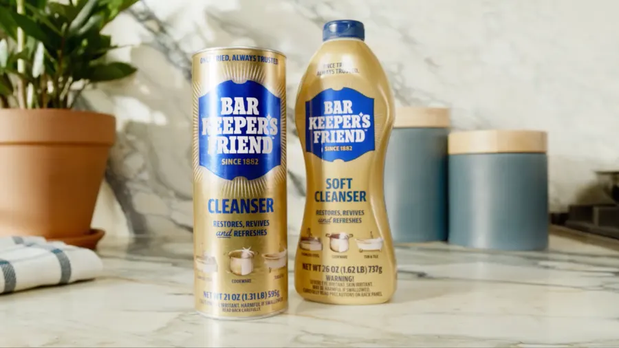 A Bar Keeper's Friend Cleanser can and a Soft Cleanser bottle standing on a marble kitchen countertop beside a potted plant and folded striped towel.