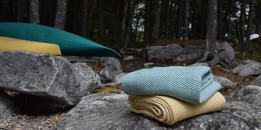 Folded teal and gold herringbone wool throws stacked on lichen-covered granite rocks beside a Maine lake, with two canoes and pine trees in the background.