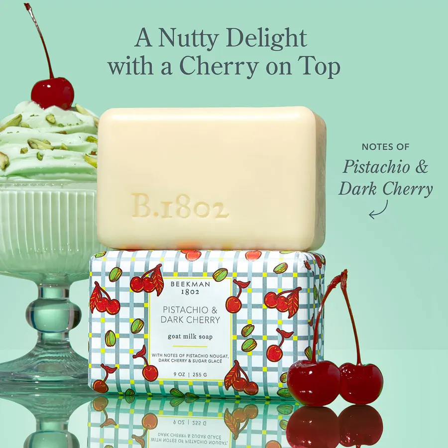 A cream-colored bar of Beekman 1802 goat milk soap embossed "B.1802" resting atop its cherry-and-pistachio patterned box, beside a mint dessert cup and fresh cherries, against a mint-green background.