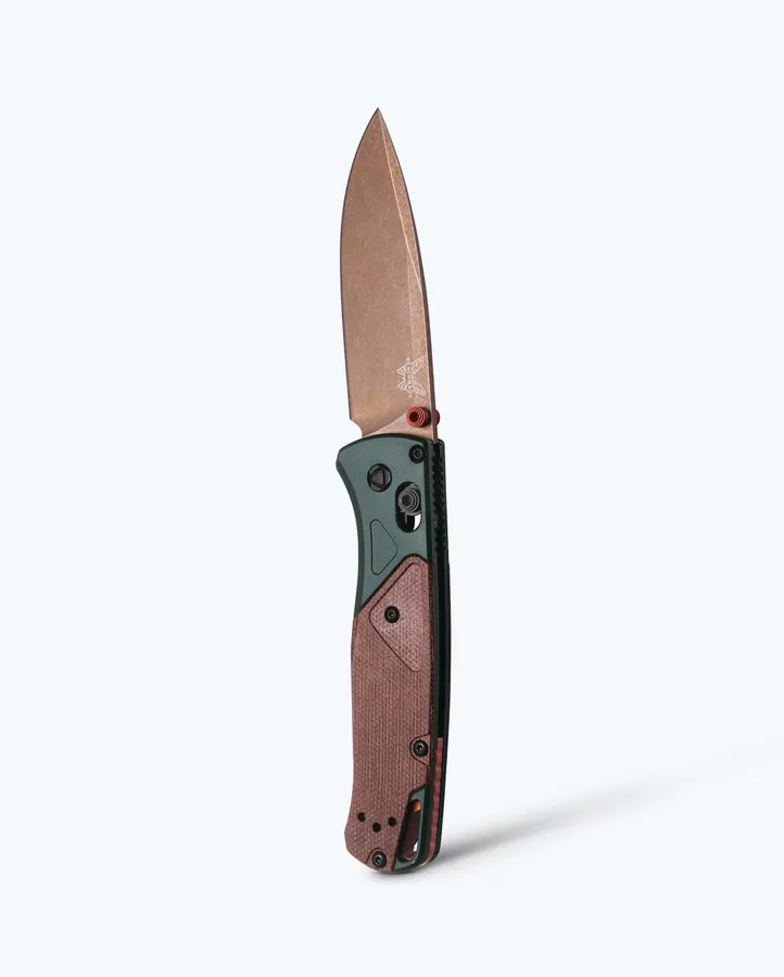 An open Benchmade Bugout folding knife standing upright, bronze drop-point blade extended, with a green anodized handle and brown canvas Micarta scales, against a plain white background.