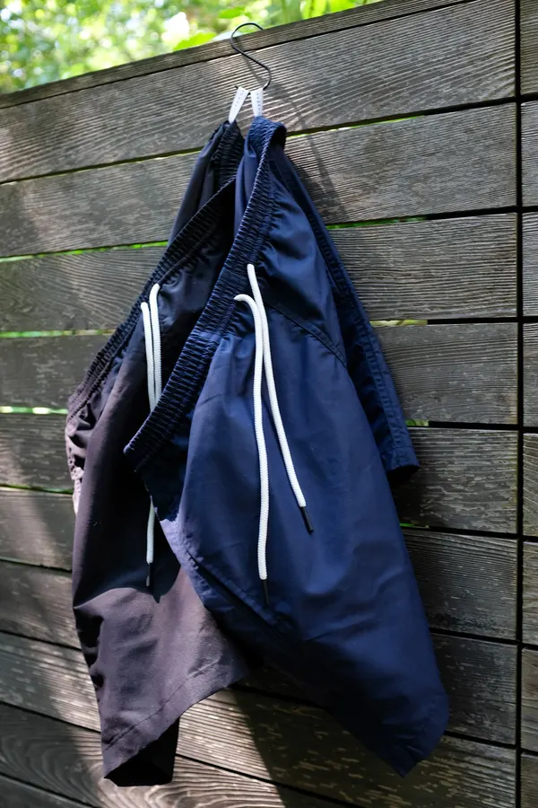 A pair of navy nylon shorts with white drawstring hanging on a wire hanger against a weathered wood-slat fence, dappled outdoor light and green foliage visible above.