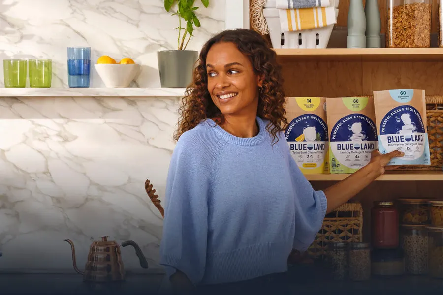 A smiling woman in a blue sweater in a kitchen, reaching for a shelf holding three Blueland refill pouches (toilet bowl, laundry, dishwasher tablets) beside houseplants and glassware.