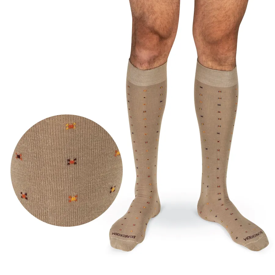A cropped view of a man's bare lower legs wearing khaki over-the-calf dress socks with a small geometric dot-and-cross pattern, with a circular macro inset of the knit pattern at lower left, on a white background.