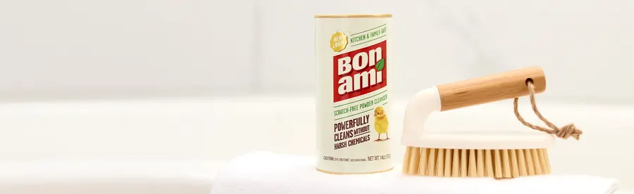 A Bon Ami powder cleanser can standing next to a wooden-handled scrub brush on the edge of a white bathtub, soft natural light.