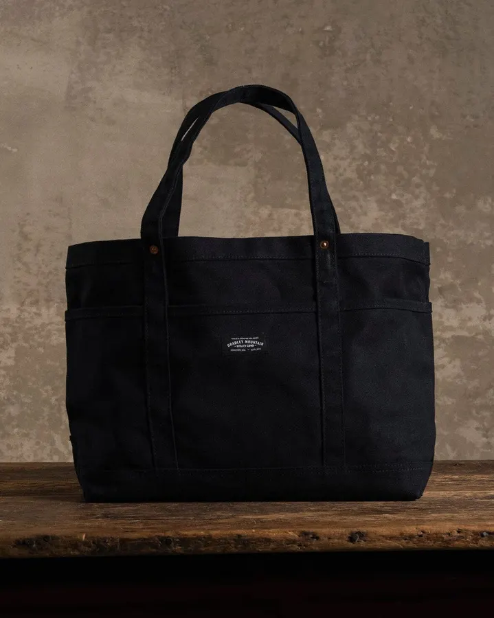 A black canvas tote bag with two handles and a front pocket bearing a small Bradley Mountain woven label, standing on a wooden table against a weathered tan wall.