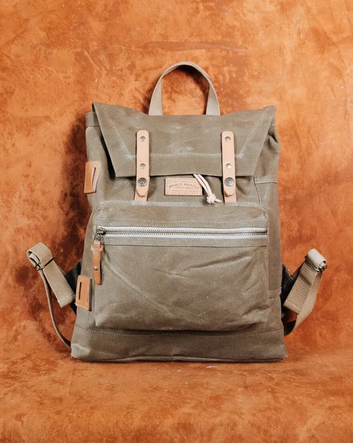A sage-green waxed canvas backpack with a roll-top flap, leather straps, and a front zip pocket, shown against a mottled rust-orange backdrop.
