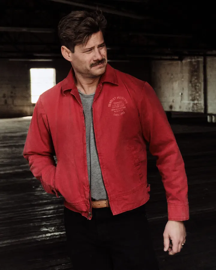 A man with a mustache wears an open red canvas work jacket over a grey t-shirt, standing in a dim, empty industrial warehouse with worn wood floors.