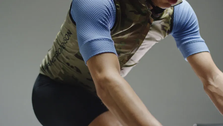 Close crop of a cyclist mid-pedal-stroke wearing a MultiCam camouflage vest with the Brandt-Sorenson wordmark down the side, over a light blue quarter-sleeve jersey, against a plain grey studio background.