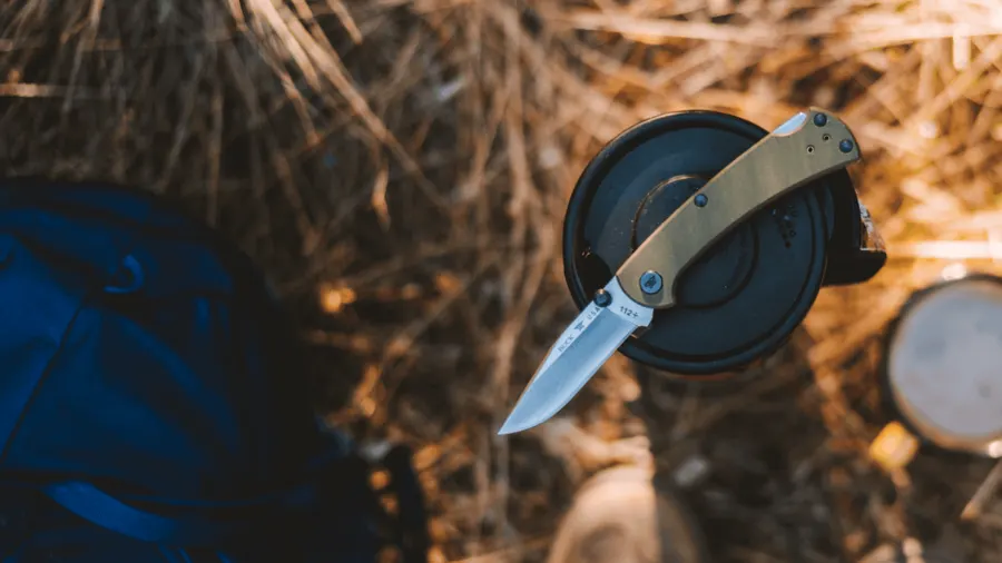 A folded Buck 112 Ranger knife with an olive-green handle balanced open atop a black camp mug, blurred dry grass and a blue pack in the background, warm late-day light.
