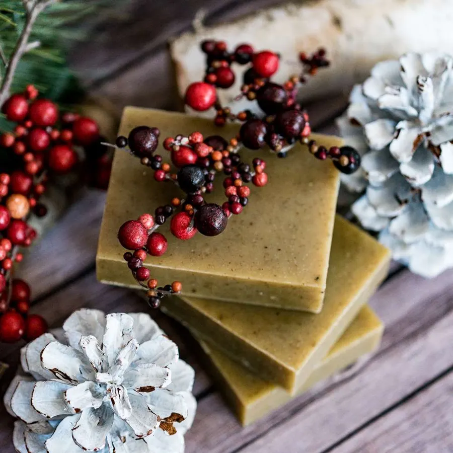 A stack of three sage-green handmade soap bars flecked with herbs, topped with a sprig of red mistletoe berries, on a wood-plank surface surrounded by white-tipped pinecones and evergreen.