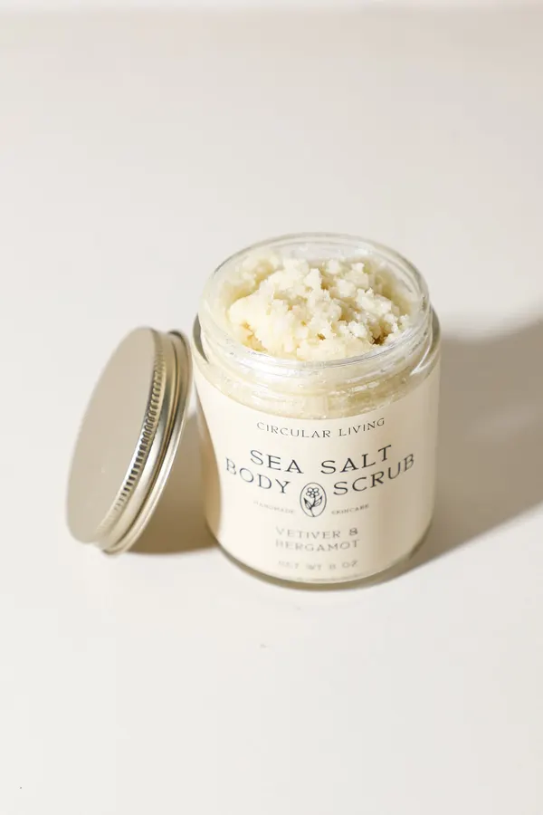 An open glass jar of Circular Living sea salt body scrub in vetiver and bergamot, its silver lid resting beside it, on a plain cream background with soft side-lit shadow.