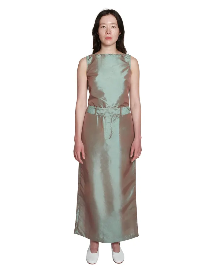 A woman in a sleeveless iridescent sage-and-bronze silk taffeta column dress with a self-belt waist, standing against a plain white background.