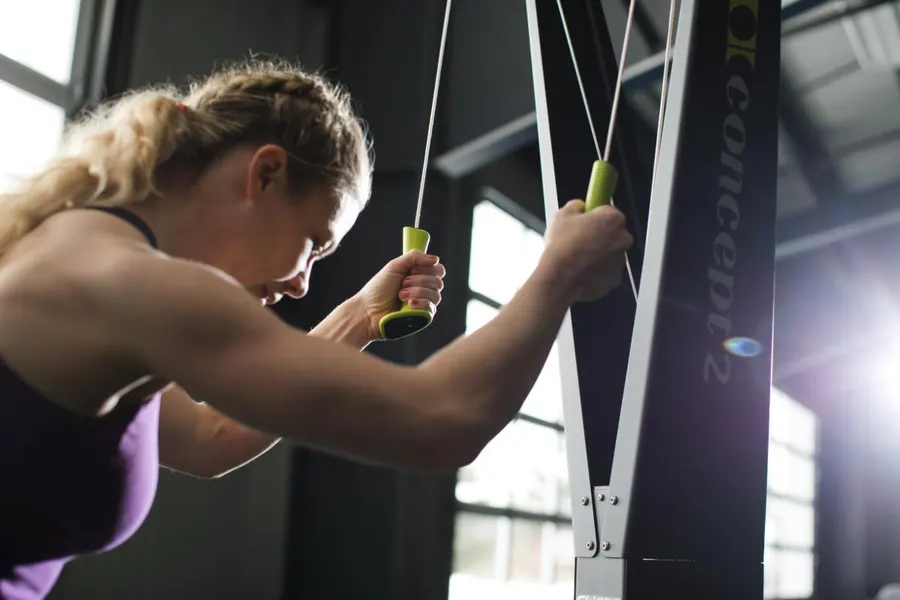 A woman doing a lunging cable-arm pull on a Concept2 machine in a gym, backlit by large windows.