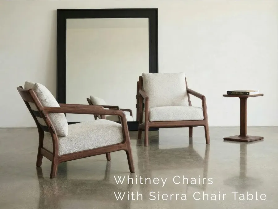 Two upholstered walnut-armed Whitney lounge chairs and a matching Sierra side table with a book on top, in front of a large black-framed mirror on a plain wall over polished concrete floor.