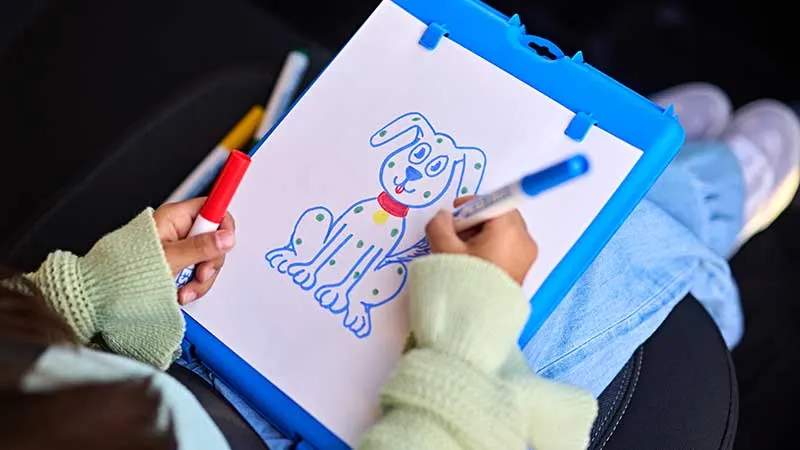 A child in a light-green sweater drawing a spotted cartoon dog on a Crayola Color Wonder art easel with a marker, seen from overhead in the back seat of a car.