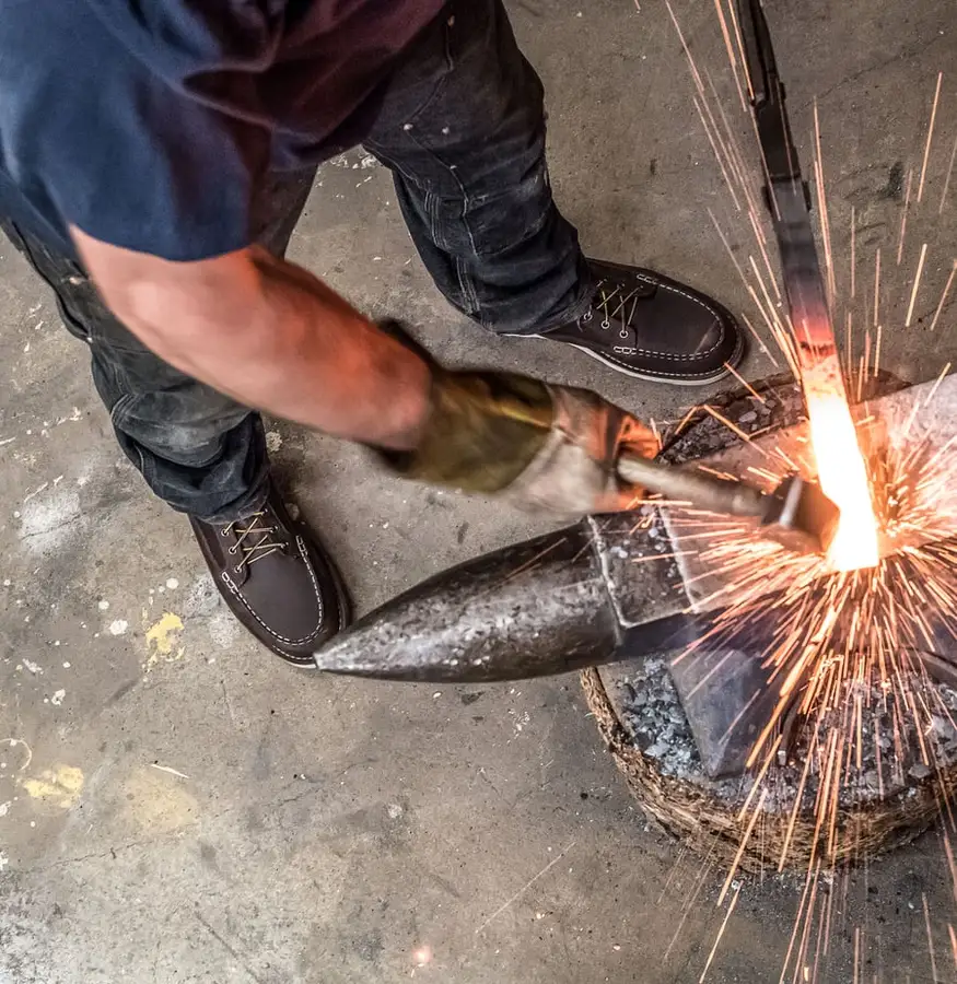 A man wearing brown leather Bull Run moc-toe work boots stands at an anvil, striking a glowing piece of metal with a hammer as sparks fly, shot from above on a concrete workshop floor.
