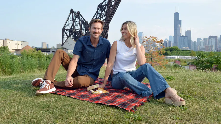 A man and a woman sitting on a red plaid picnic blanket in a grassy lot with the Chicago skyline and a raised railroad bridge behind them, both wearing Dearborn Denim jeans.