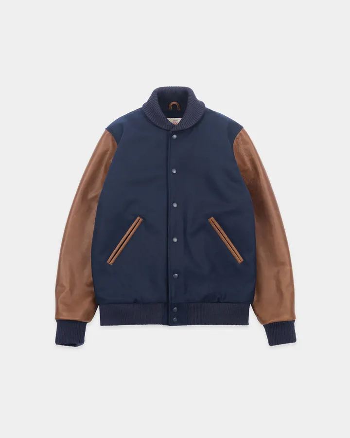 A navy wool varsity jacket with brown leather sleeves and knit collar, cuffs and waistband, laid flat on a plain light-grey background.