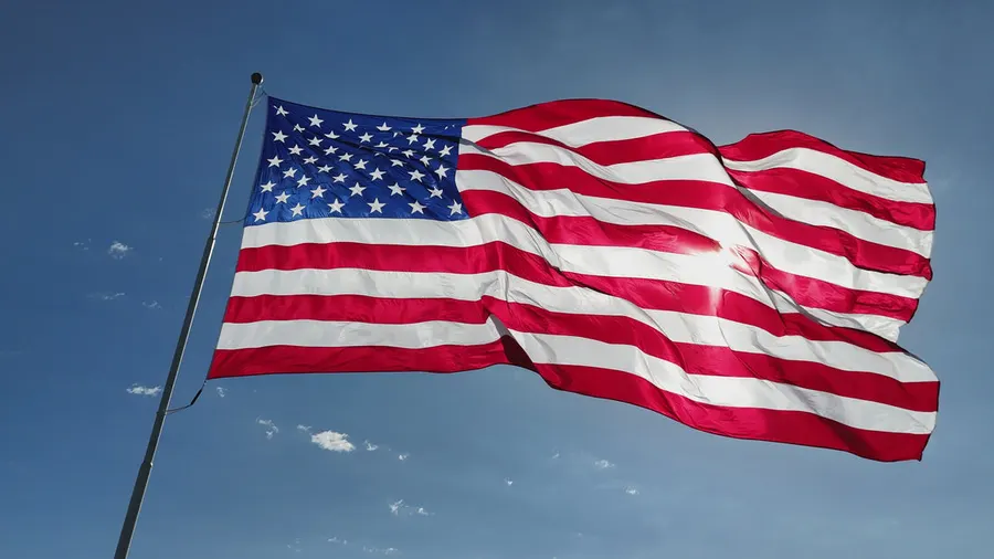 A large American flag with full stars and stripes flies from a pole against a mostly clear blue sky, backlit by the sun.