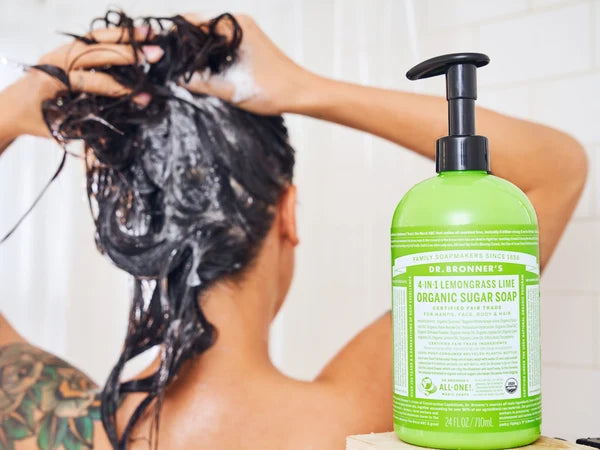 A woman seen from behind lathers shampoo into wet dark hair in a shower; a green Dr. Bronner's 4-in-1 Organic Sugar Soap pump bottle sits on the tub ledge beside her.