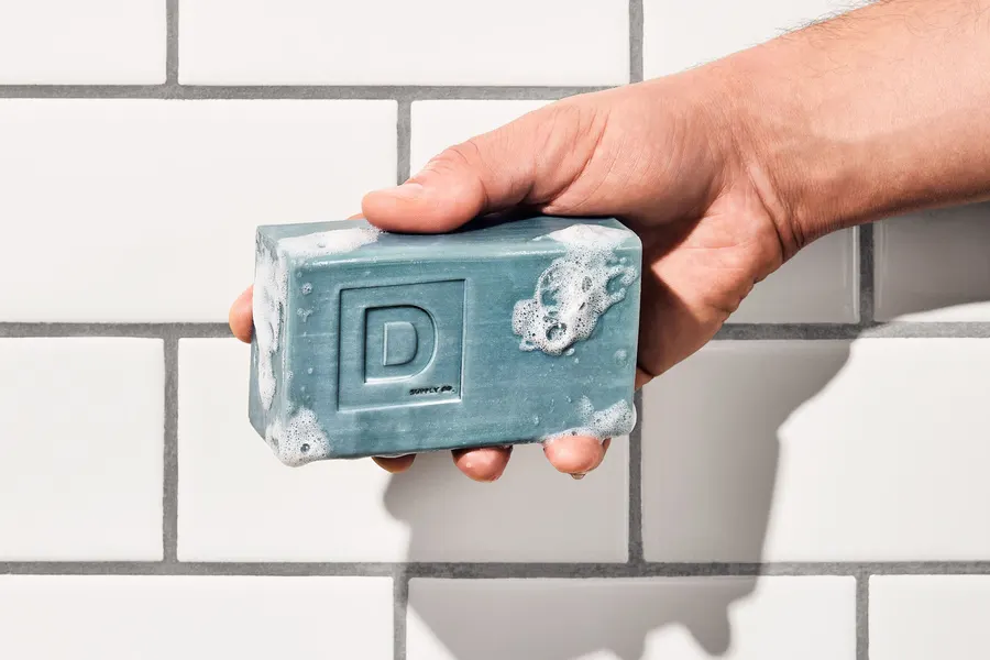 A man's hand holds up a sudsy grey-blue bar of soap embossed with a large letter D, against a white subway-tile shower wall.