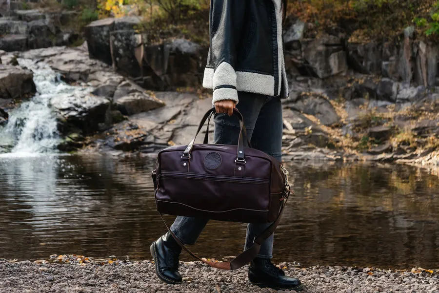 A person in dark boots and jeans carries a burgundy leather duffel bag with handles, walking along a rocky shoreline beside a small waterfall.