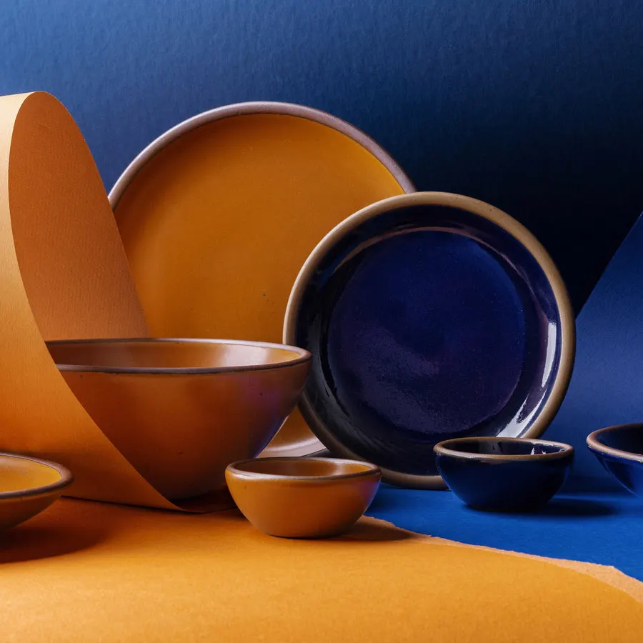 A colorful studio still life of East Fork stoneware: a mustard-yellow plate and bowl and a cobalt-blue plate with small bowls, arranged against a bold blue-and-orange paper backdrop.