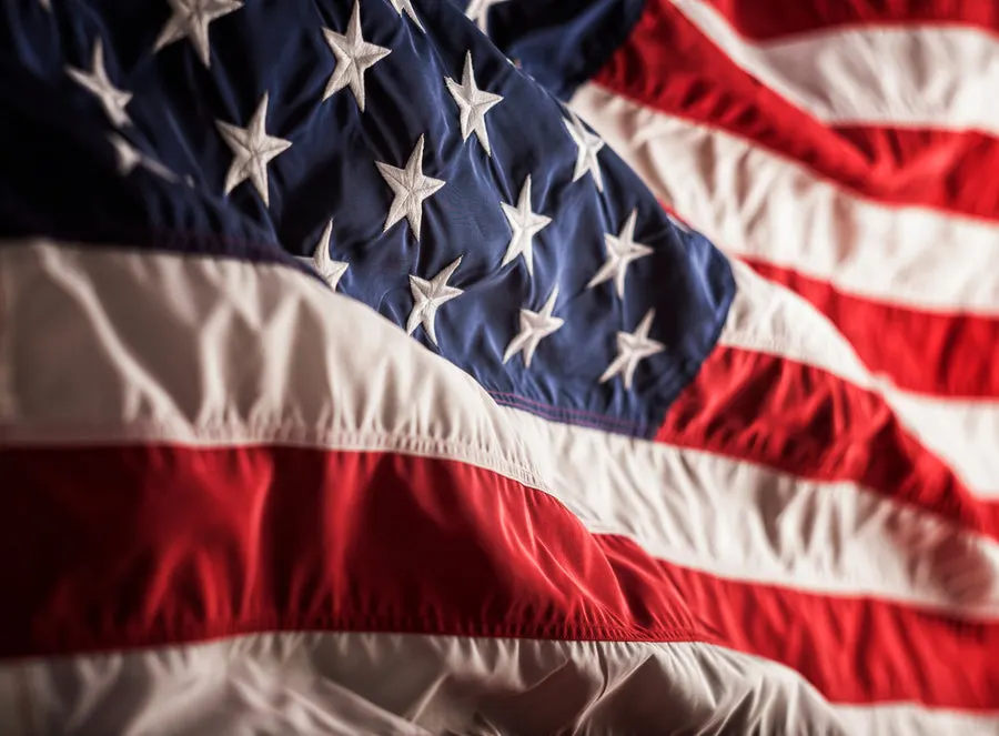 A close-up of an American flag's embroidered white stars on blue field and sewn red-and-white stripes, rippling with warm directional light.