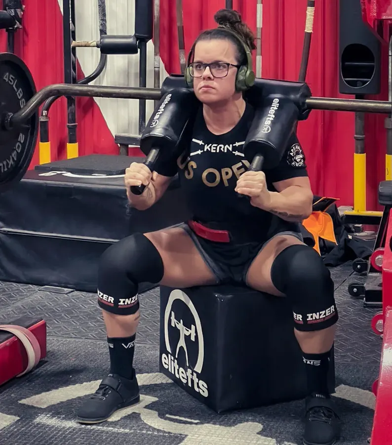 An athlete wearing knee sleeves sits on a black elitefts squat box in a gym, gripping an SS Yoke Bar resting across padded shoulder supports, red curtains and rack in the background.