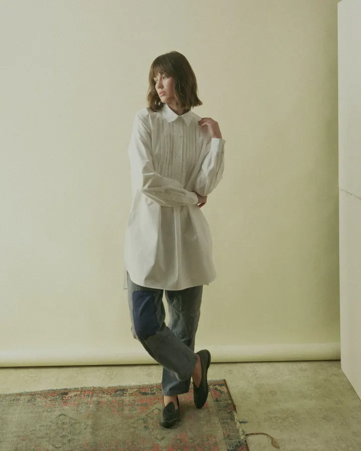 A woman in an oversized white pintucked poplin shirt and grey trousers stands on a patterned rug against a cream wall, looking off to the side.