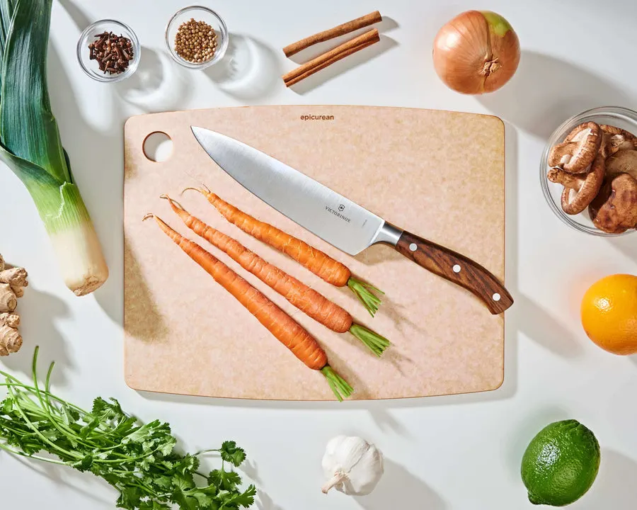 Overhead view of a natural-colored Epicurean cutting board on a kitchen counter with a chef's knife and whole carrots on top, surrounded by leeks, onion, garlic, citrus, and mushrooms.