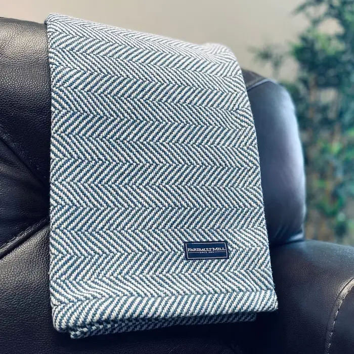 A blue-and-white herringbone cotton throw with a Faribault Mill label, draped over the back of a black leather chair with a potted plant blurred in the background.