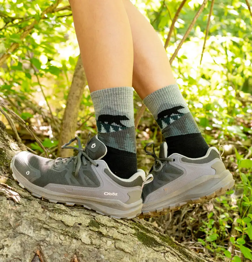 A hiker's bare lower legs wearing gray crew socks with a black bear and pine-tree motif, paired with gray trail running shoes, stepping on a mossy log in sunlit woods.